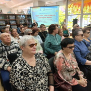 На переднем плане группа читателей НОСБ, сидящих в читальном зале, по центру на заднем плане виден экран с текстом: «Для тех, чью осень не покинула весна»